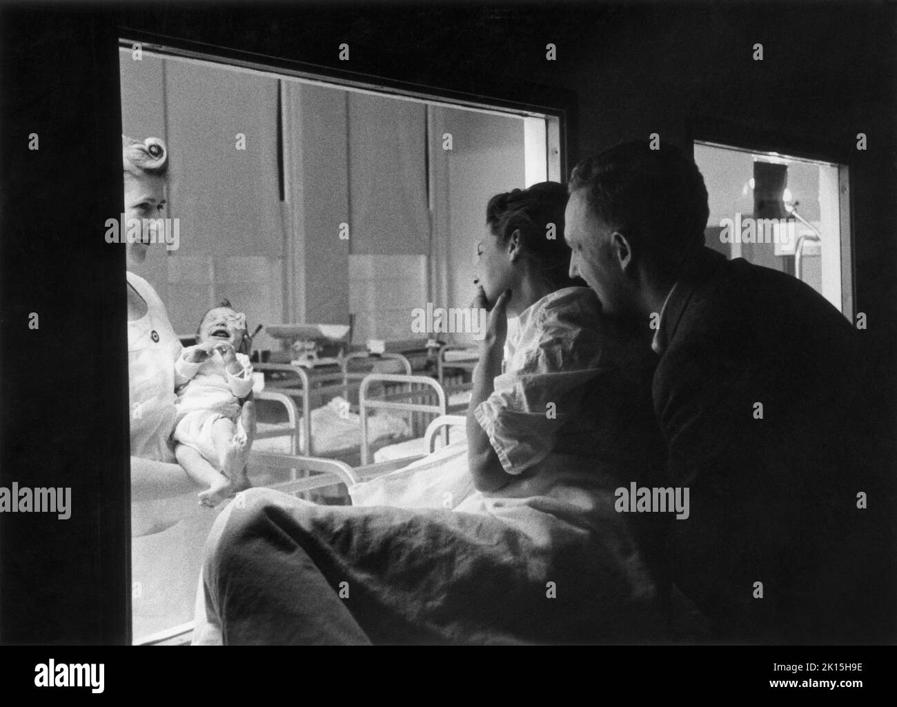 Couple regarding their new baby through the window of a hospital nursery. Stock Photo