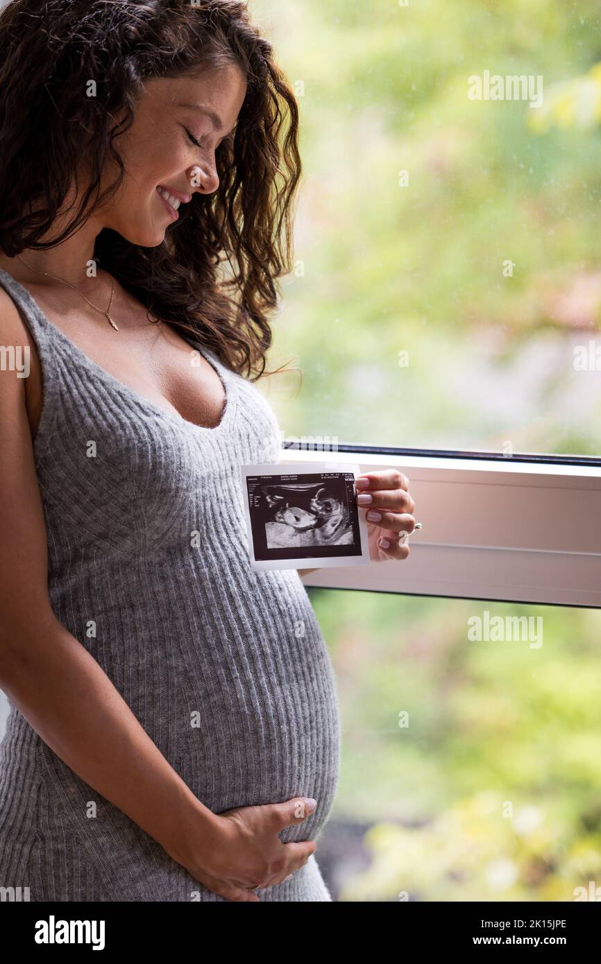 Pretty handsome pregnant woman holding belly and looking at ultrasound image of baby in front of window at home Stock Photo