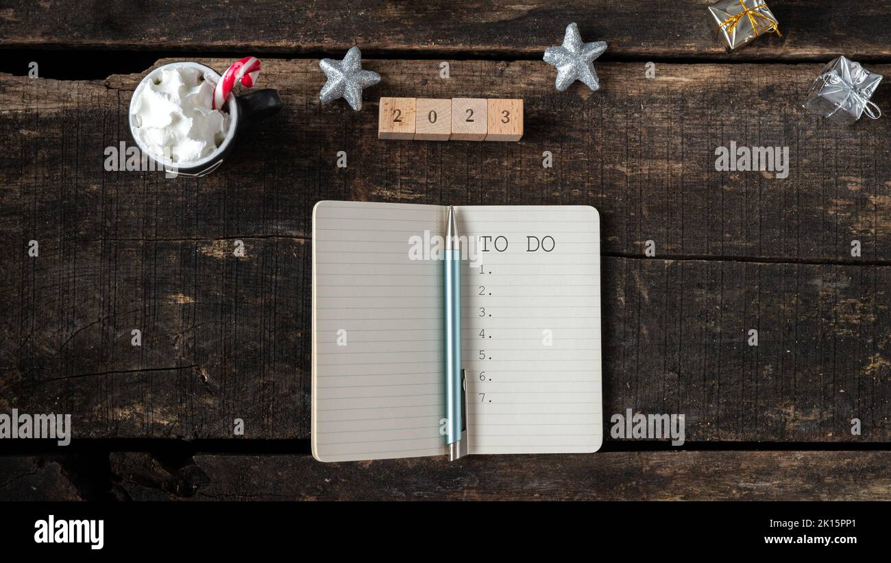 Top view of 2023 sin on wooden blocks next to a To do list in a notepad and holiday hot cocoa drink placed on wooden desk decorated with christmas orn Stock Photo