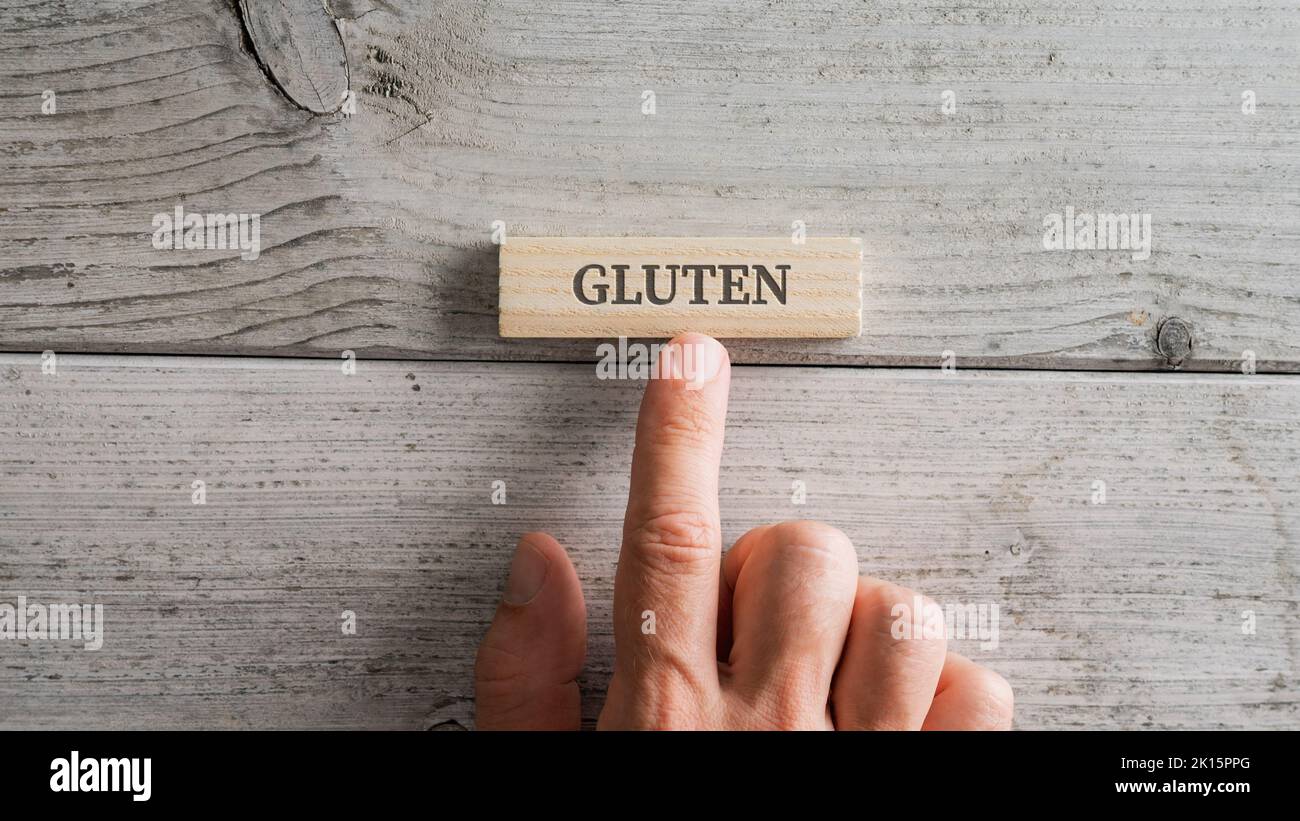 Male hand placing wooden peg with a Gluten sign on it over white textured wooden background. Stock Photo