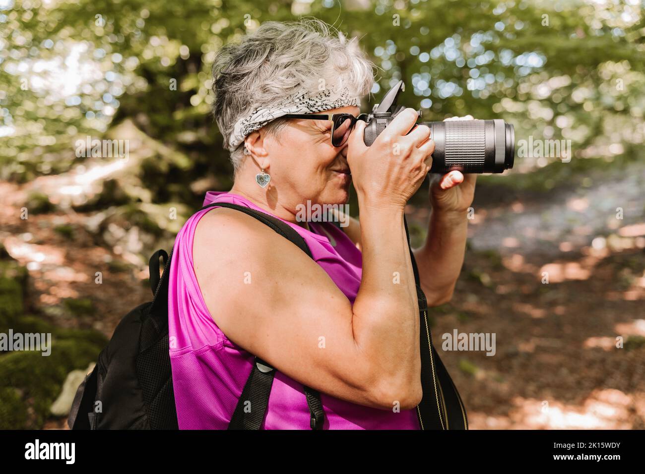 Side view of elderly female tourist with backpack shooting nature while spending sunny summer day in park Stock Photo