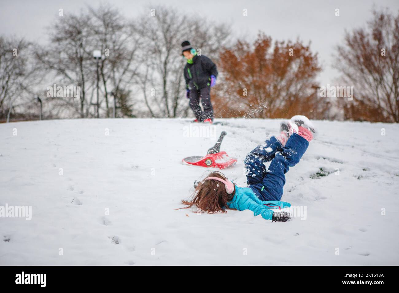 A little girl falls off sled while boy snowboards behind her Stock