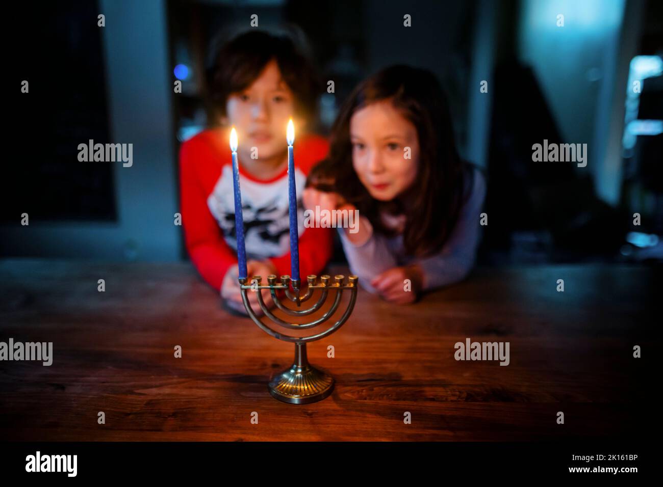 Two siblings sit at table together staring at lit candles on menorah Stock Photo
