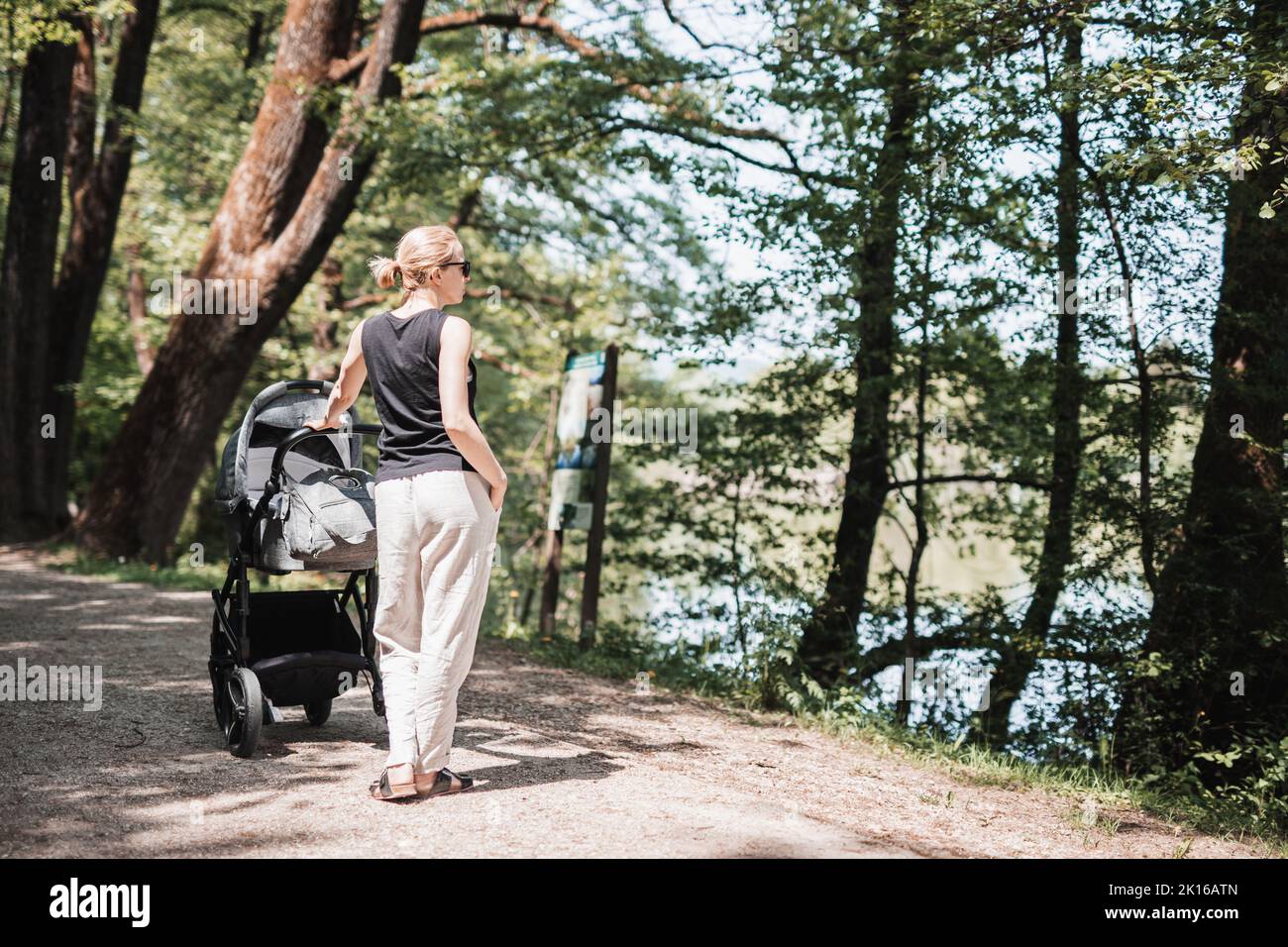 Rear view of casualy dressed mother walking with baby stroller in city park. Family, child and parenthood concept. Stock Photo