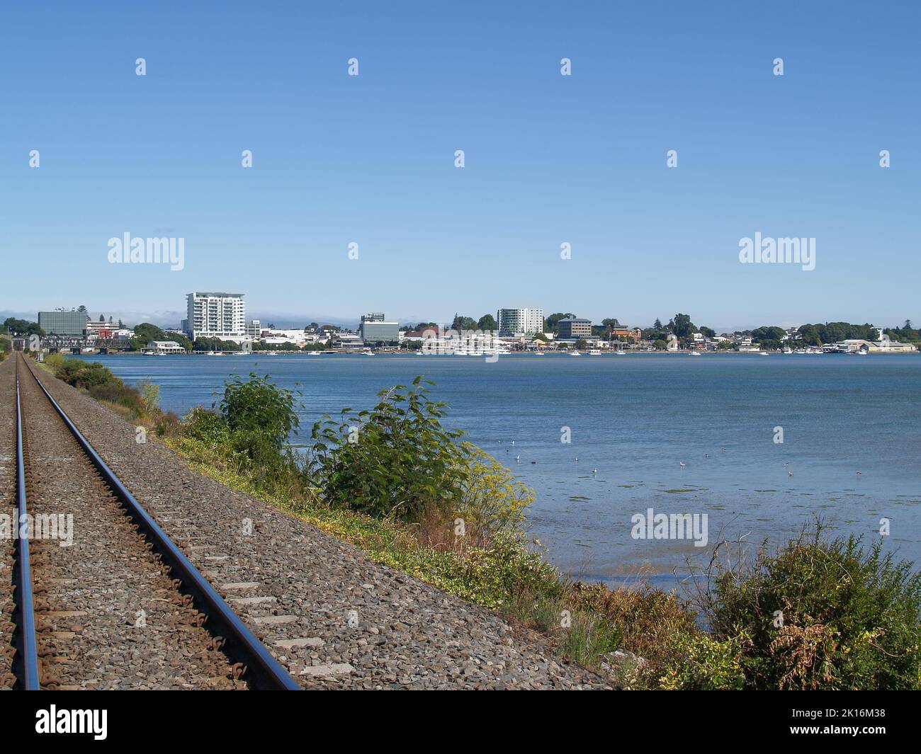Distant view of Tauranga business district buildings and harbour edge following leading lines of railway track across harbour. Stock Photo