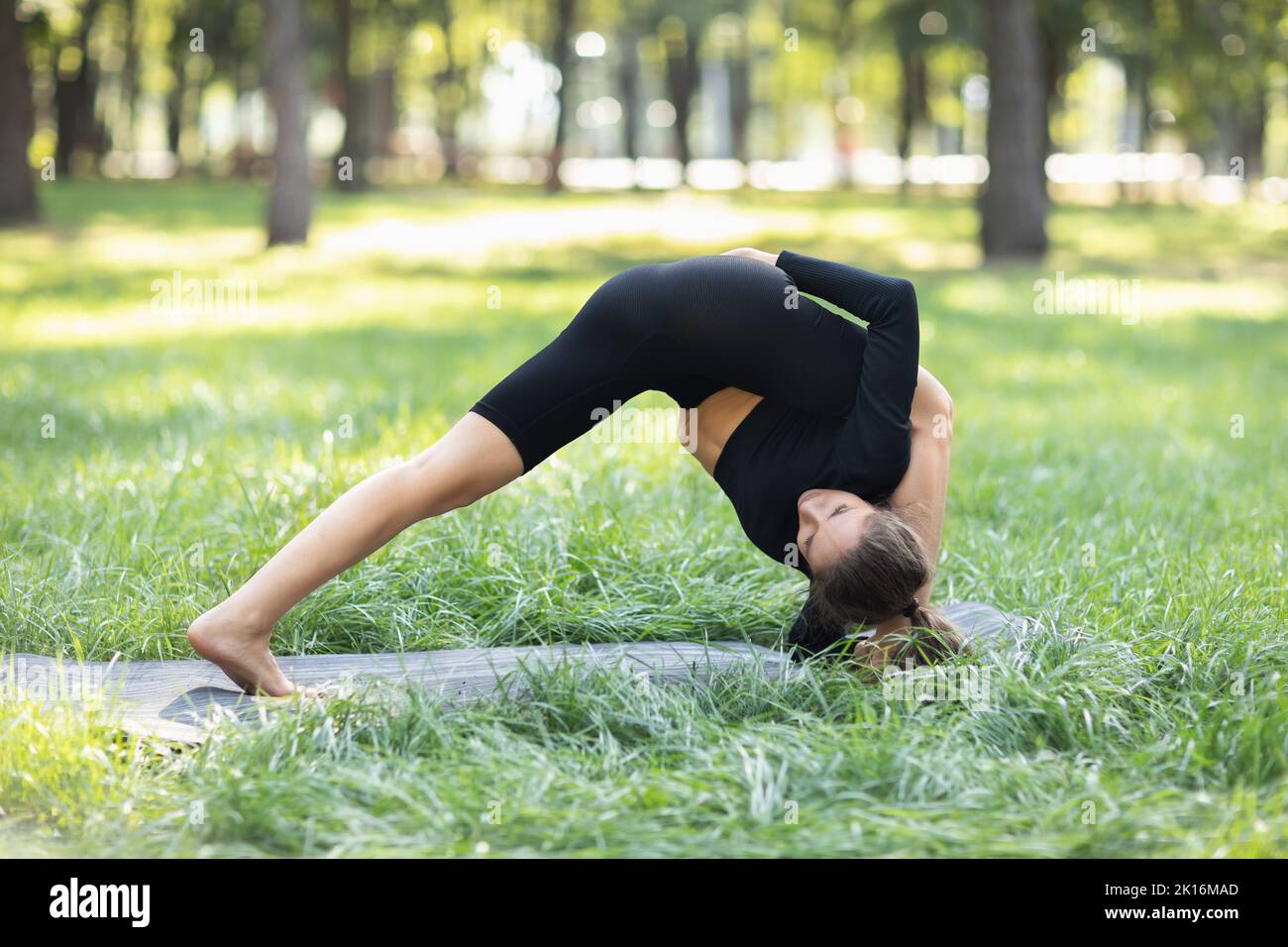 Young woman in black sportswear practicing yoga doing lead exercise to Ruchikasana, exercising on a summer morning in the park, standing on a mat Stock Photo
