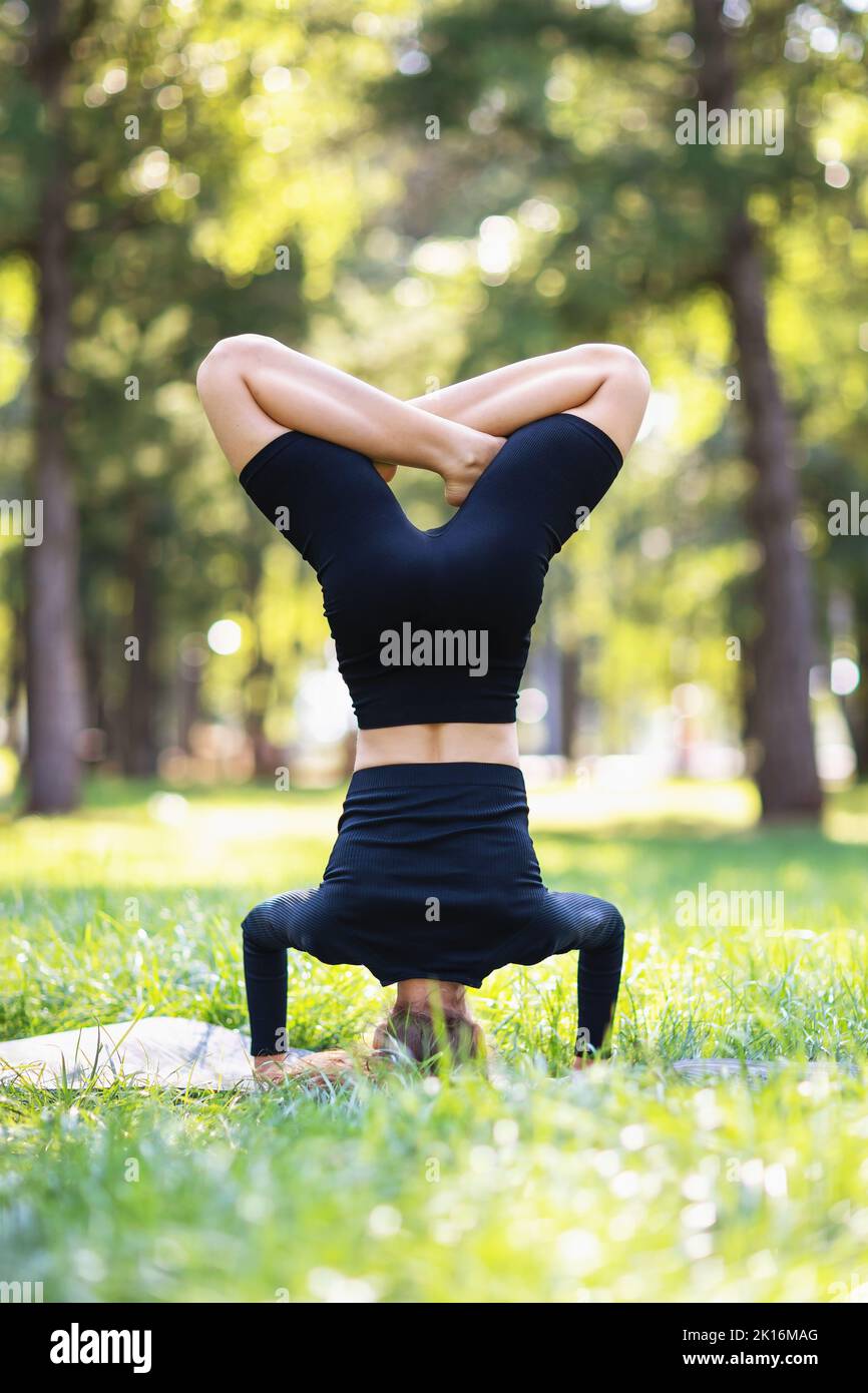 Woman in black sportswear practicing yoga doing a variation of shirshasana exercise, headstand with lotus, exercising in the park on a warm summer mor Stock Photo