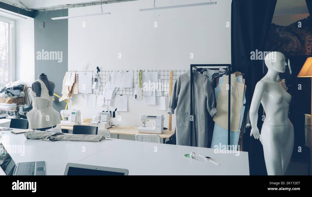 Light clothing design studio with large tailor's desk, mannequins, numerous sketches pinned on wall, sewing machines, tailoring items and half-finished garments on rails. Stock Photo