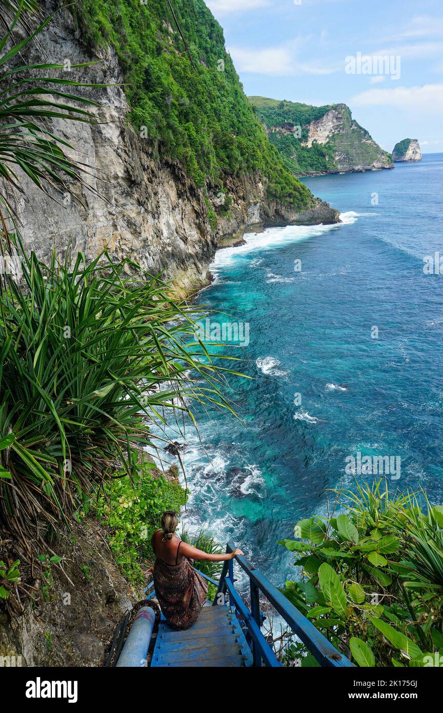 Girl standing on blue steps looking over the cliff to the crashing waves of the ocean in Nusa Penida, Bali, Indonesia. Stock Photo