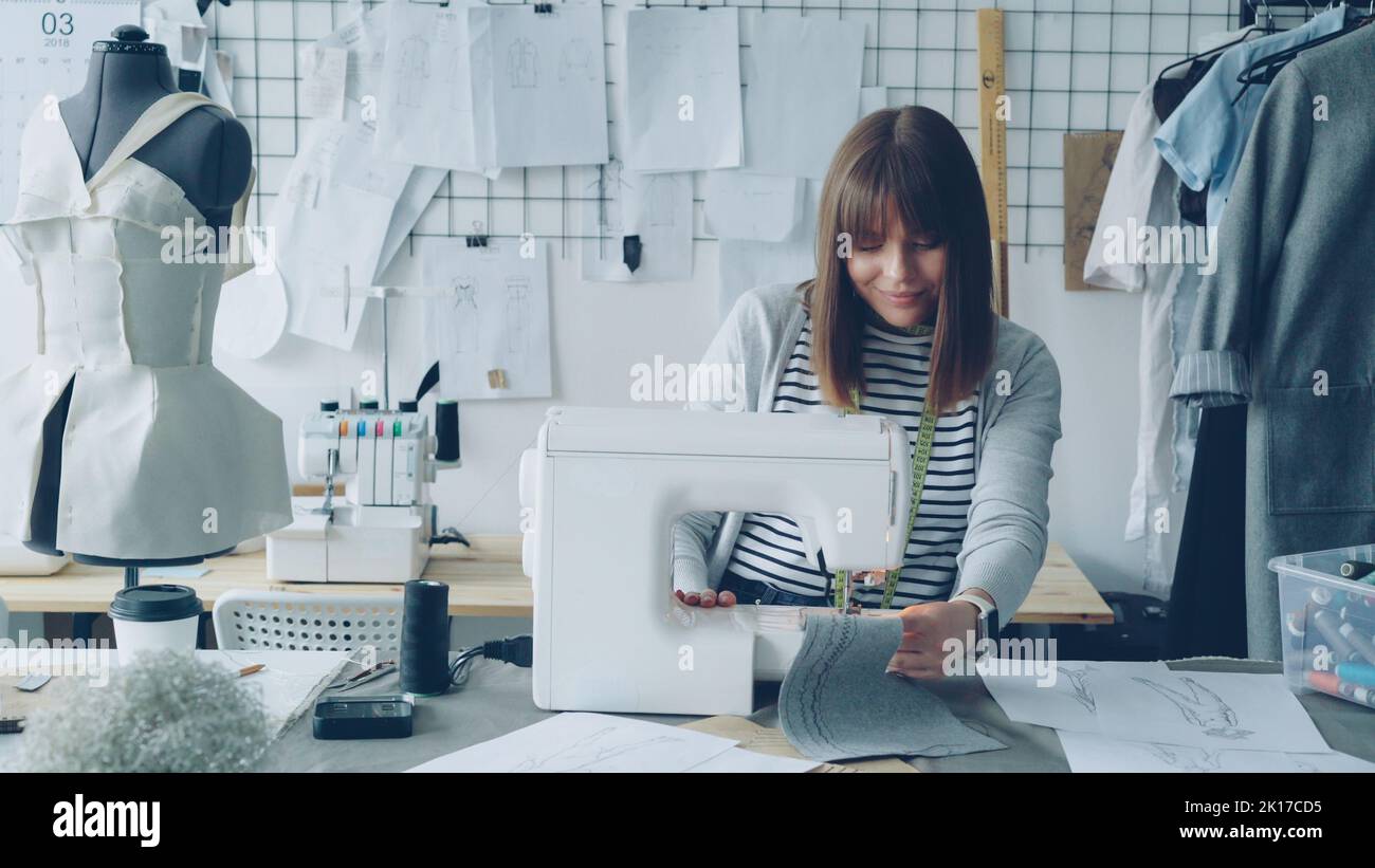 Young attractive dressmaker is working on sewing machine and looking at ...