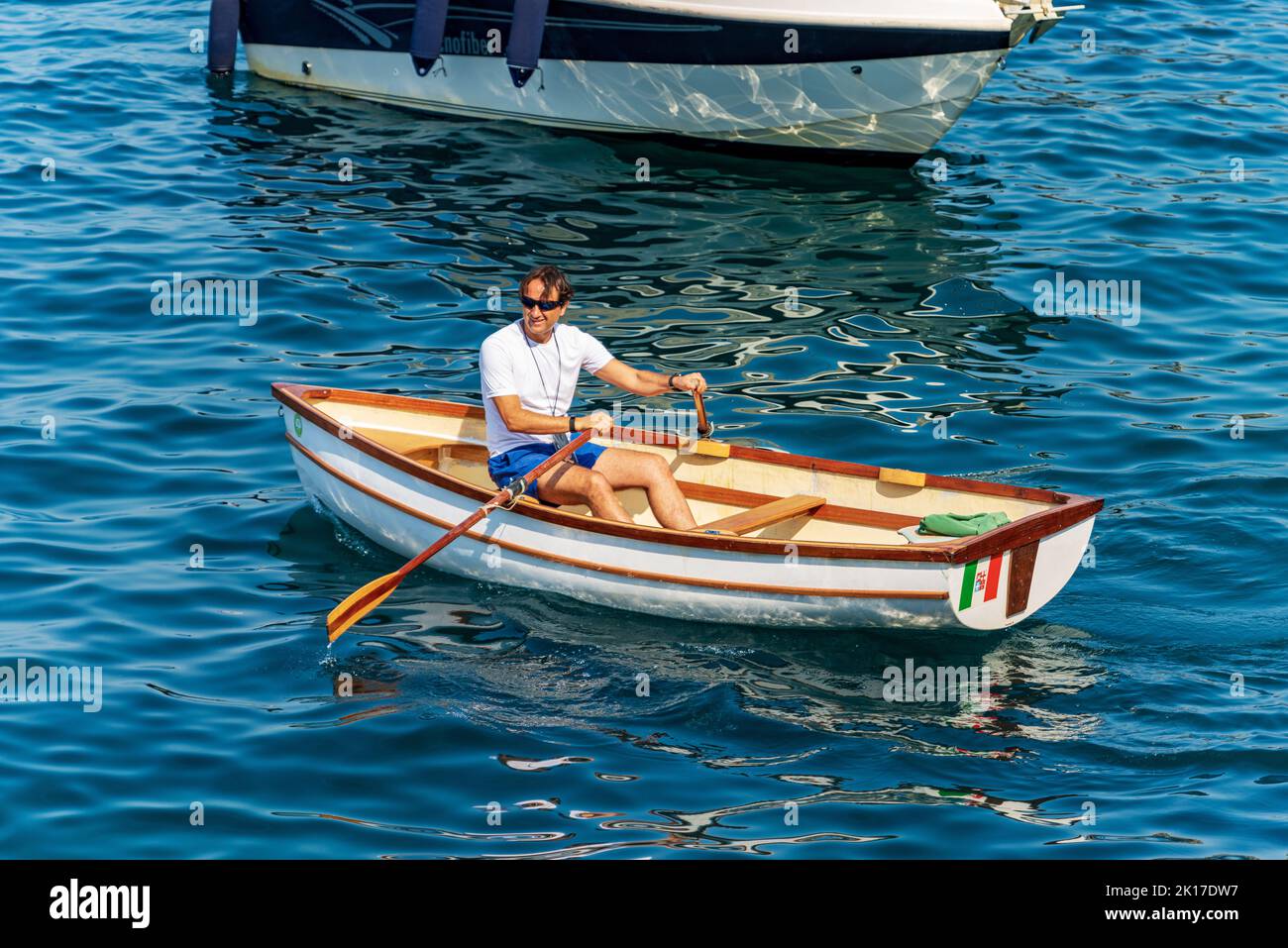 Adult man aboard of a small white and brown wooden rowing boat in the blue Mediterranean sea in front of the ancient Tellaro village, Liguria, Italy. Stock Photo