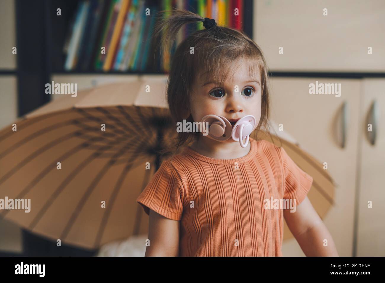 Funny portrait of a baby girl standing in the house with two pacifiers in her mouth looking to one side. Happy infant. Family people indoor Interior Stock Photo