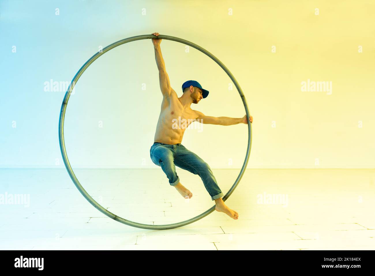 Full body of shirtless barefoot male acrobat performing stunt with cyr wheel during training against light background in modern studio Stock Photo