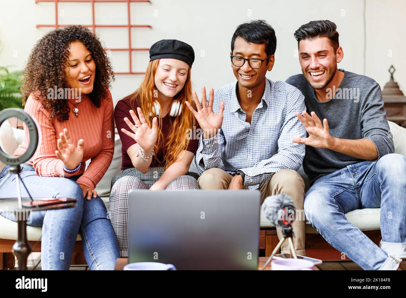 Group of positive multiracial friends waving hands while having video call via laptop on table with microphone and ring lamp Stock Photo