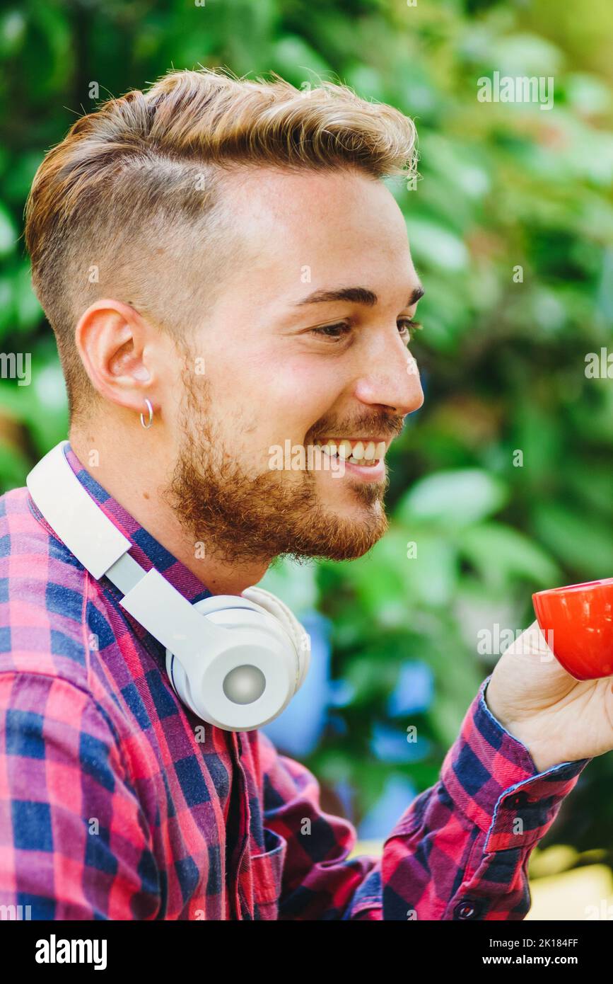 Side view of positive bearded male with modern wireless headphones drinking hot espresso while sitting on terrace of outdoor cafeteria Stock Photo