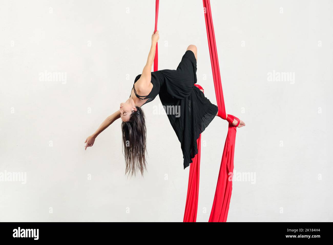 Full body barefoot woman in black dress closing eyes and bending back while hanging on red aerial silks against gray background Stock Photo