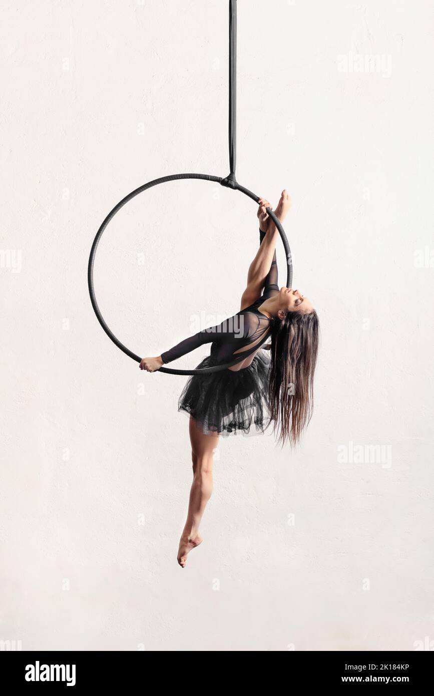 Full body barefoot dancer with long dark hair doing split pose on aerial ring during performance against gray background Stock Photo