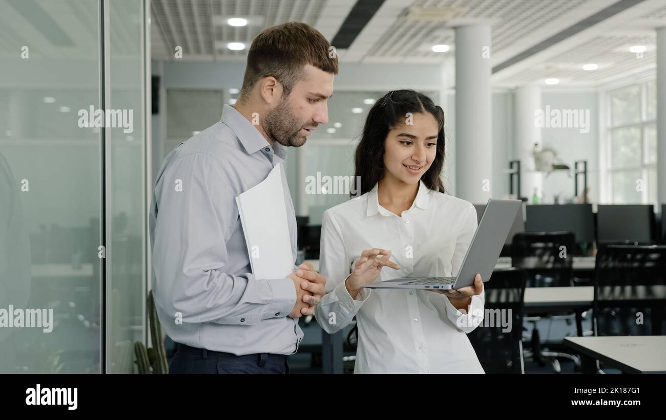 Positive friendly colleagues chatting in office discussing project details young female employee consults with male mentor shows presentation on Stock Photo