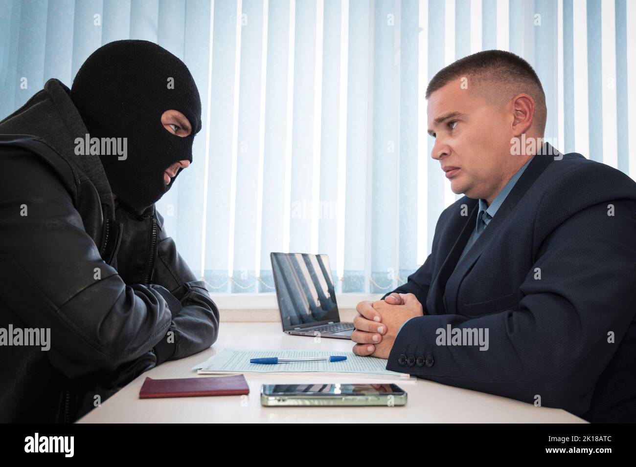 a businessman and robbers are sitting at a table. A racketeer in a black balaclava forces to sign a contract. The concept of a raider takeover of the Stock Photo