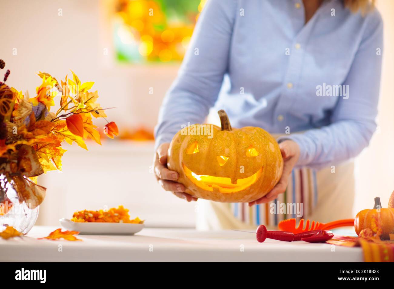Woman carving pumpkin for Halloween celebration. Female in apron cutting jack o lantern for traditional trick or treat decoration. Decorated living ro Stock Photo