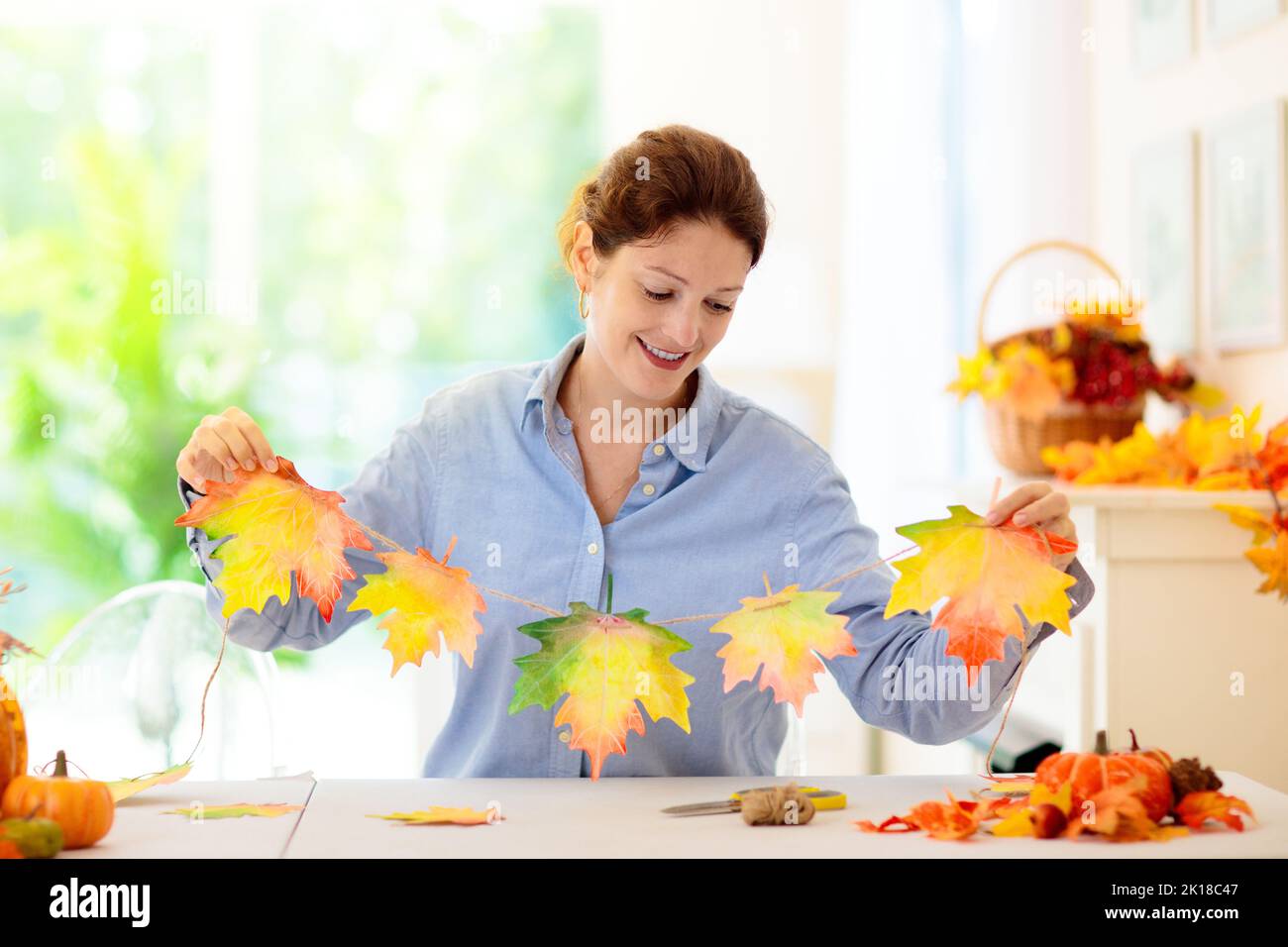 Autumn home decoration. Family decorating house with colorful maple leaves banner. Thanksgiving or Halloween celebration in white living room. Woman w Stock Photo