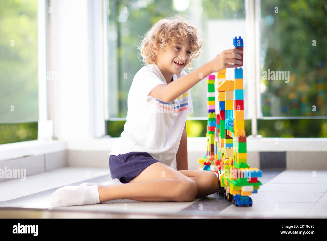 Child playing on home balcony. Outdoor patio of city apartment building. Safe glass railing for little kids. Boy building tower on sunny terrace. Stock Photo