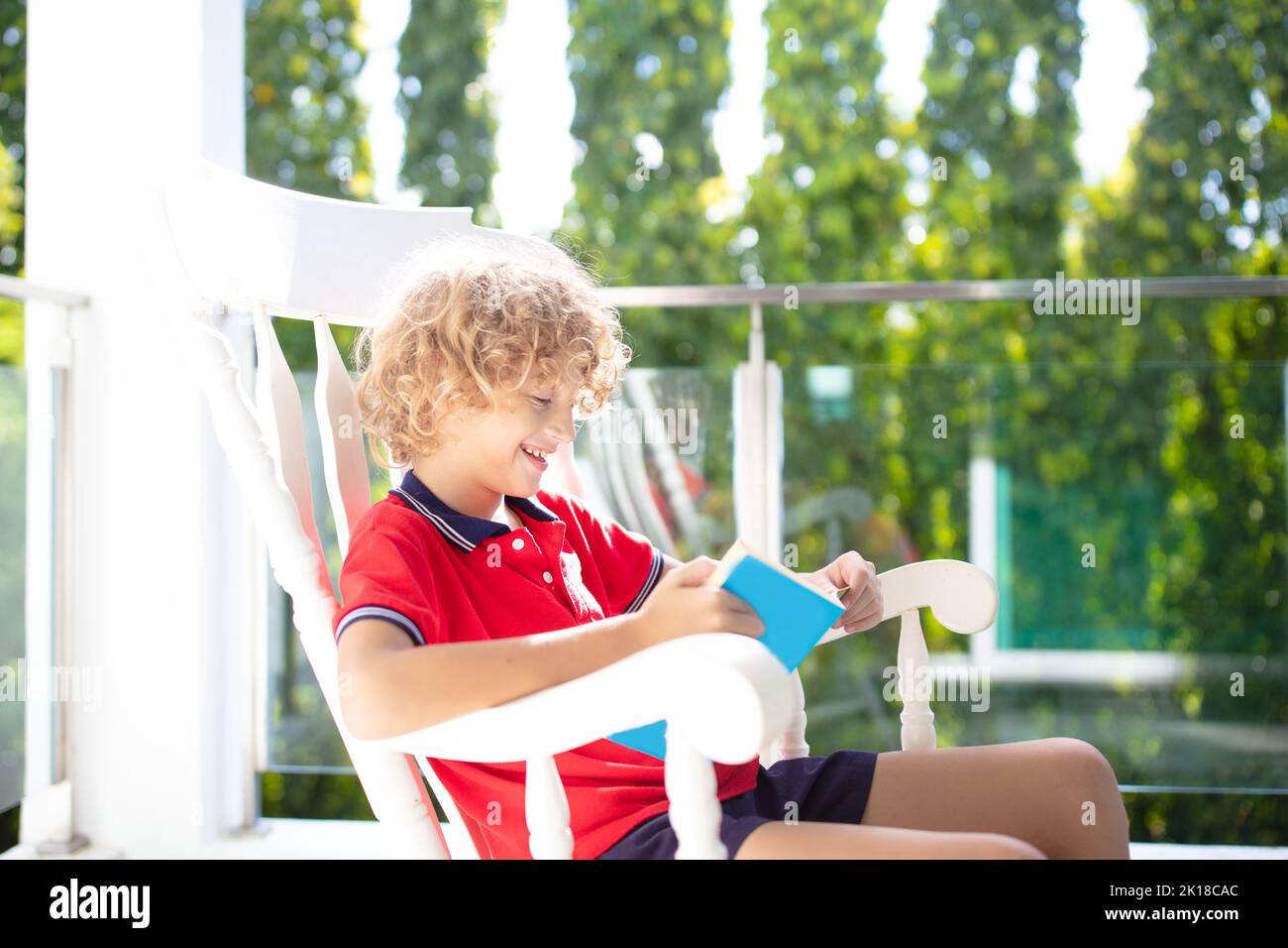 Child reading on home balcony. Outdoor patio of city apartment building. Safe glass railing for little kids. Boy playing on sunny terrace. Stock Photo