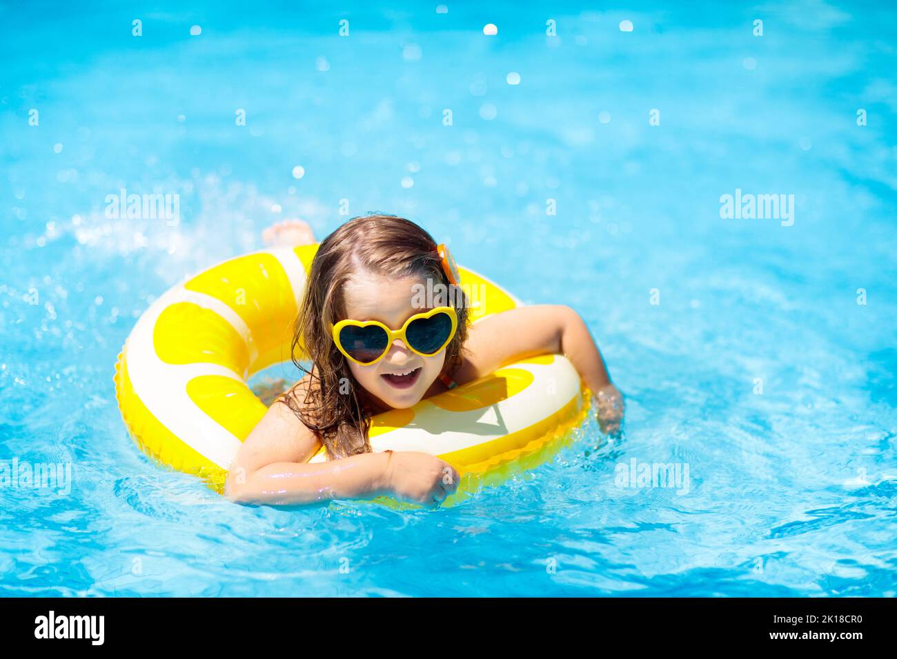 Child in swimming pool on inflatable yellow lemon ring. Little girl learning to swim with orange float. Water toy for baby and toddler. Healthy outdoo Stock Photo