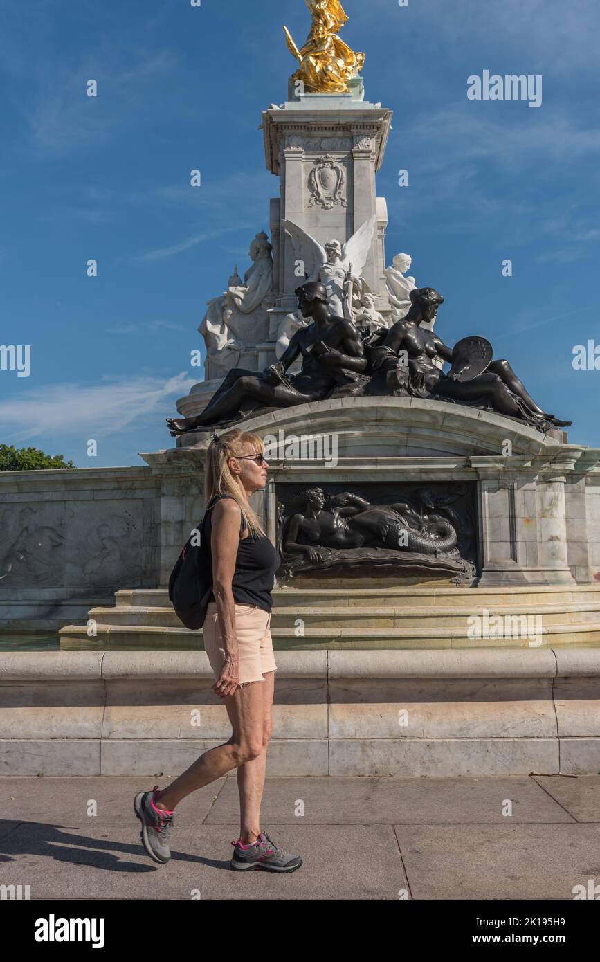Mature blonde woman with glasses and backpack walking in front of Queen Victoria monument in view of Buckingham Palace. Stock Photo