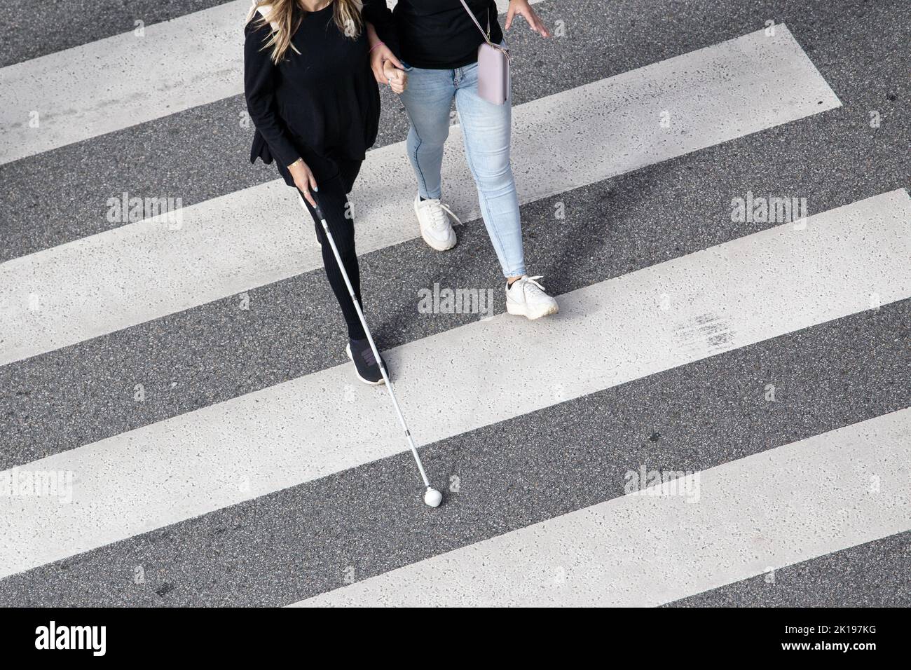 Scene of a Blind woman walking on zebra crossing helped by another person using her white cane. Help in the early stages of blindness Stock Photo