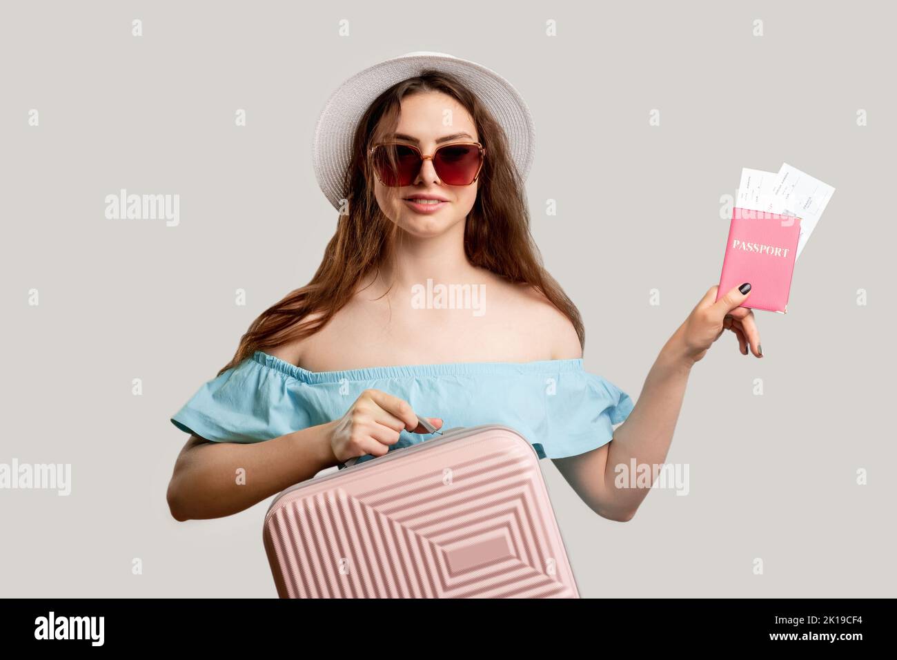 Summer holiday. Flight check-in. Happy woman in vacation outfit holding pink suitcase and foreign id boarding pass. Looking at camera isolated on neut Stock Photo