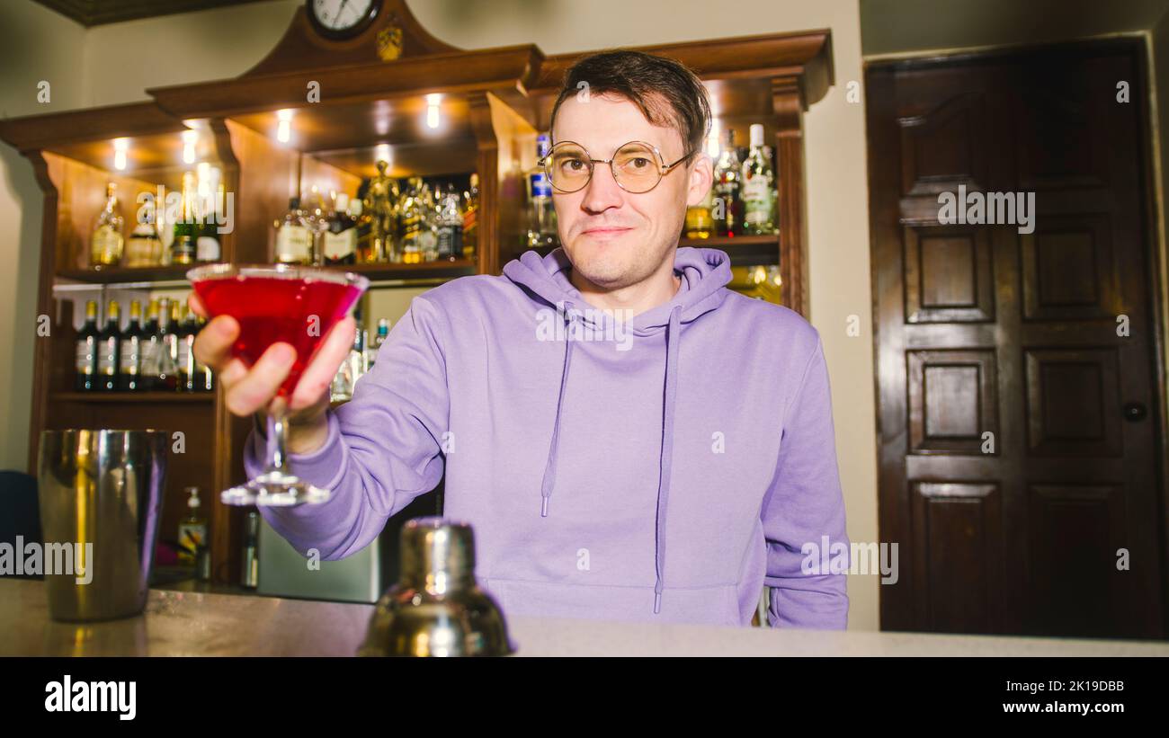 Male bartender in eyeglasses with cocktail standing behind bar, looking at camera, offering alcoholic beverage, smiling Stock Photo