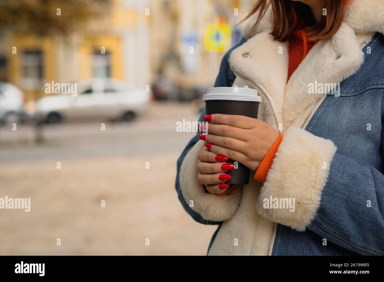 Paper cup with coffee in hands of unrecognizable woman, standing in city street. Body part of female in warm clothes with bright manicure holding hot Stock Photo