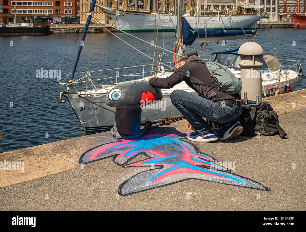 Europe, France, Dunkerque - July 9, 2022: Male artist paints pink-blue-black fish image on bollard standing at Port du Bassin de la Marine quay. Museu Stock Photo