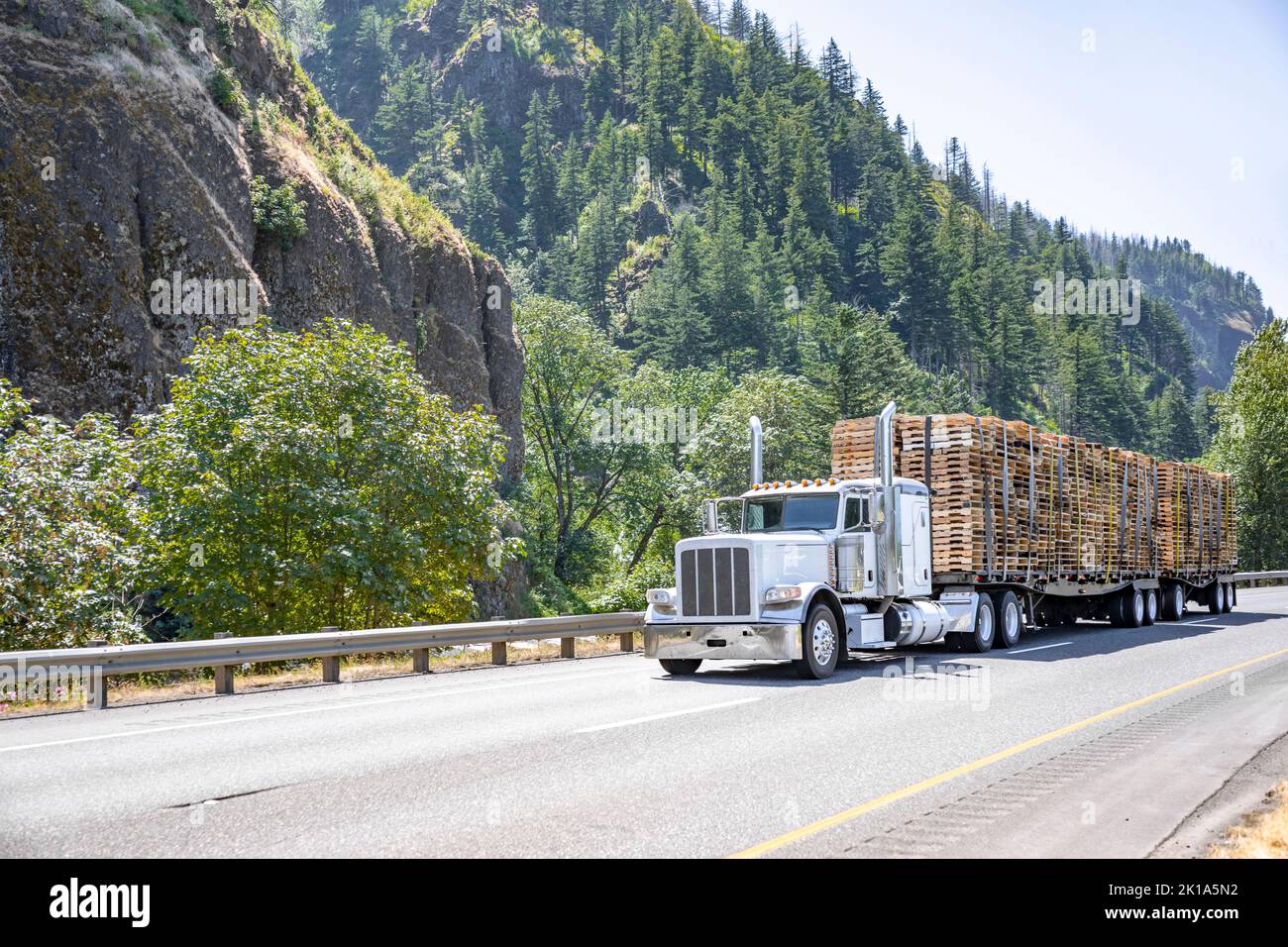 Classic style white big rig semi truck tractor with extended cab transporting fastened wood pallets on flat bed semi trailers running on the wide divi Stock Photo