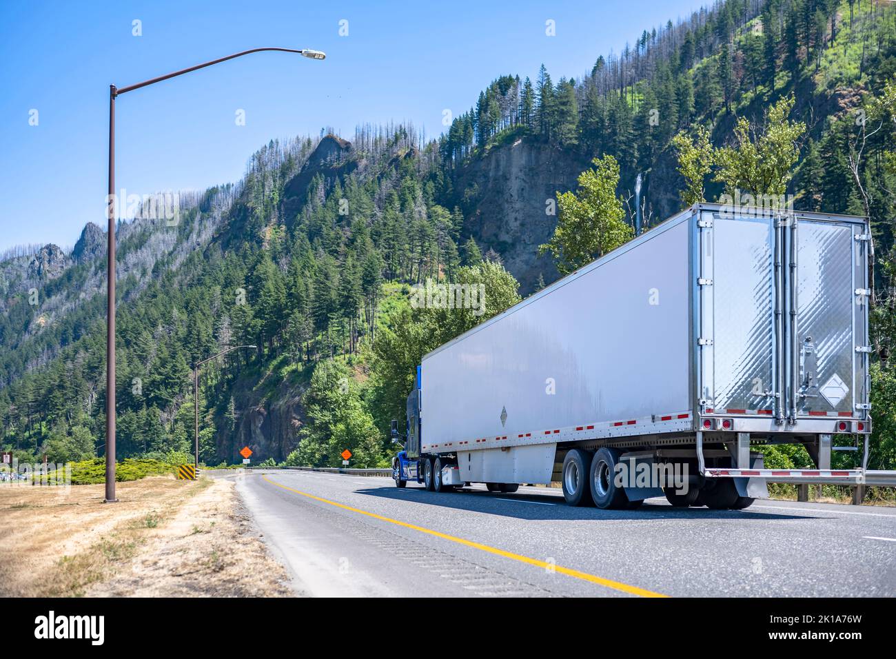 Classic blue long haul big rig semi truck tractor with chrome exhaust pipes transporting commercial cargo in refrigerator semi trailer moving on the h Stock Photo