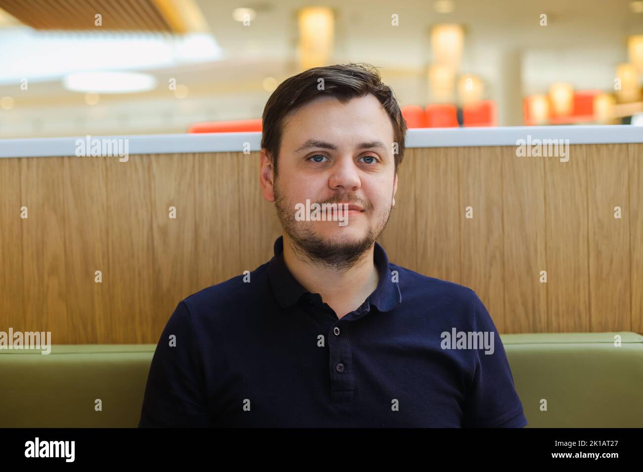 looking at the camera smiling young 32 years old caucasian unshaven beard man in a navy blue polo shirt in cafe at the food court. Waiting concept, he Stock Photo