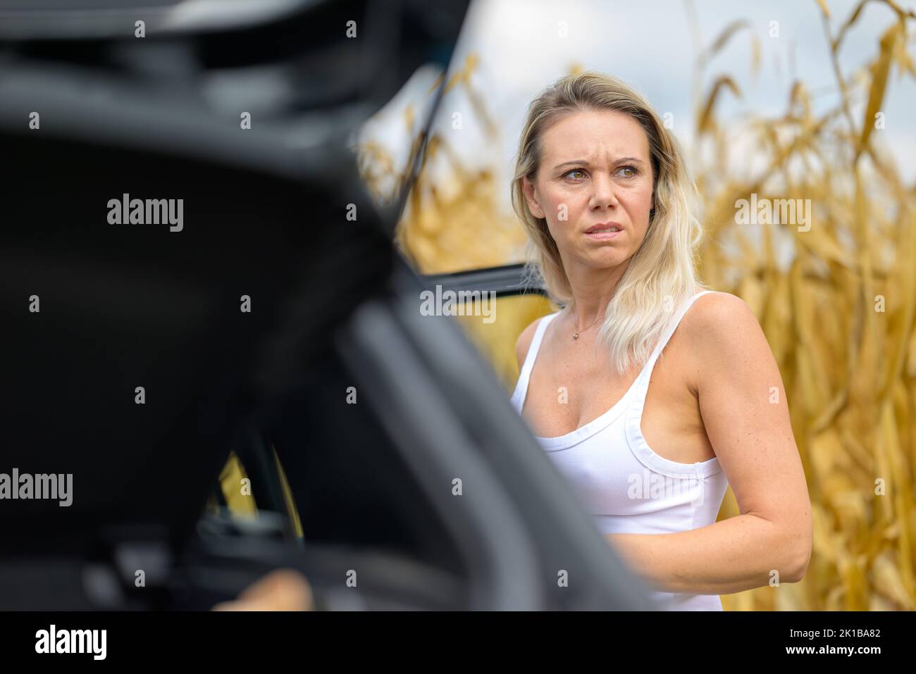Woman standing by a car with a thoughtful look because of an car failure in front of a cornfield Stock Photo