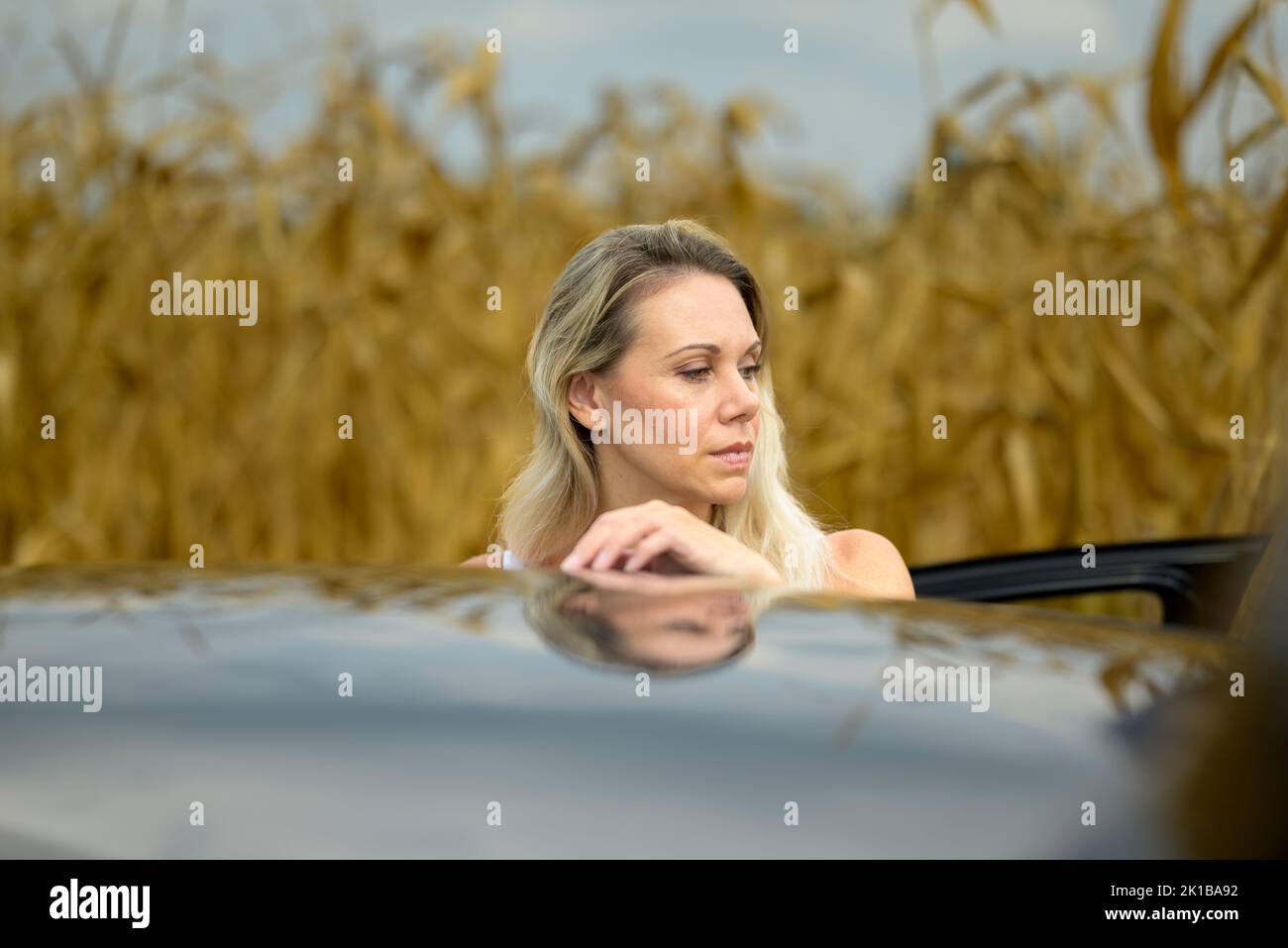 Woman standing by a car with a thoughtful looking aside in the background a cornfield Stock Photo