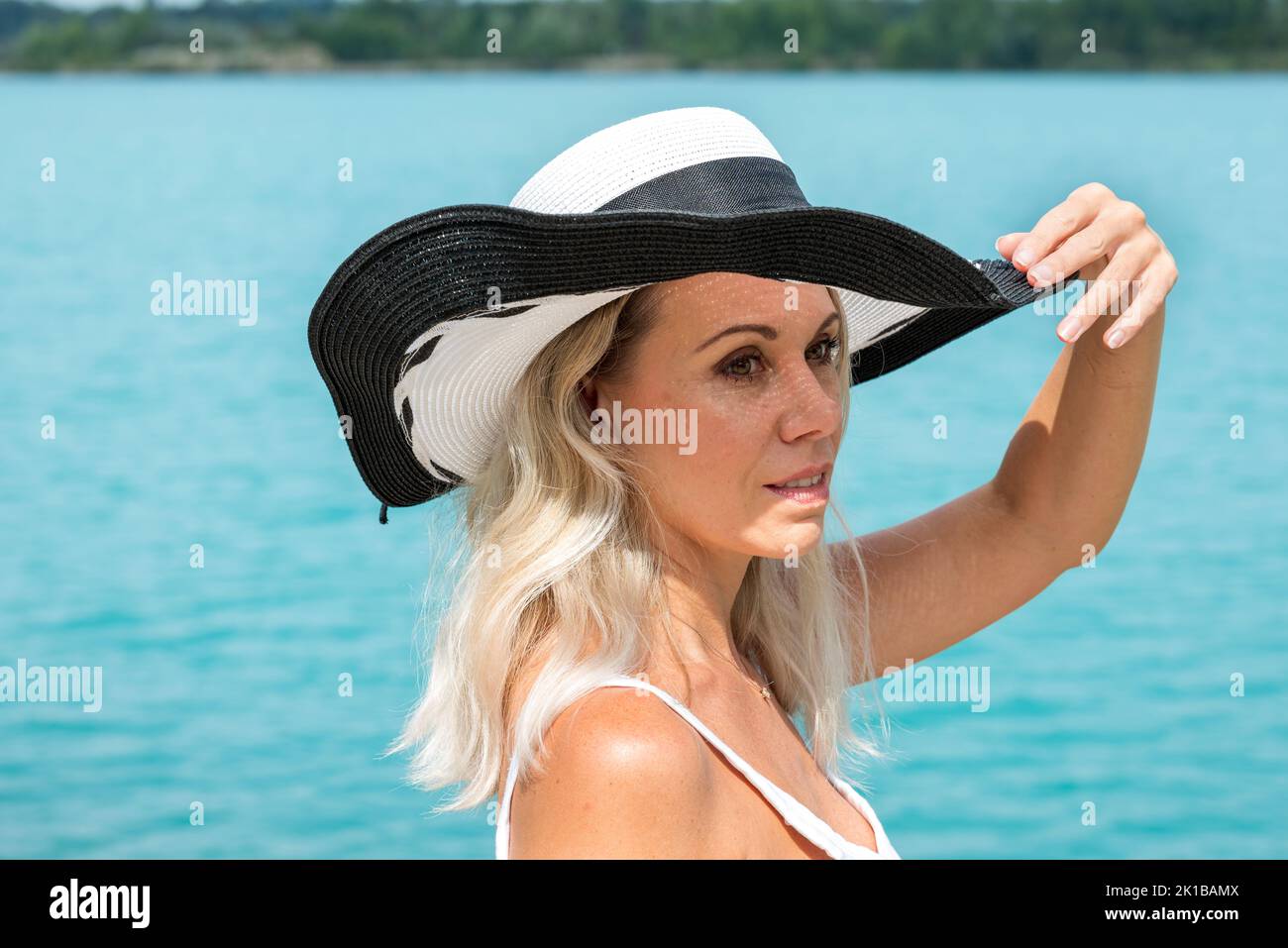 Attractive blond woman smiling friendly wearing a black and white summer hat in a white top with her hands on the hat in front of a turquoise sea look Stock Photo