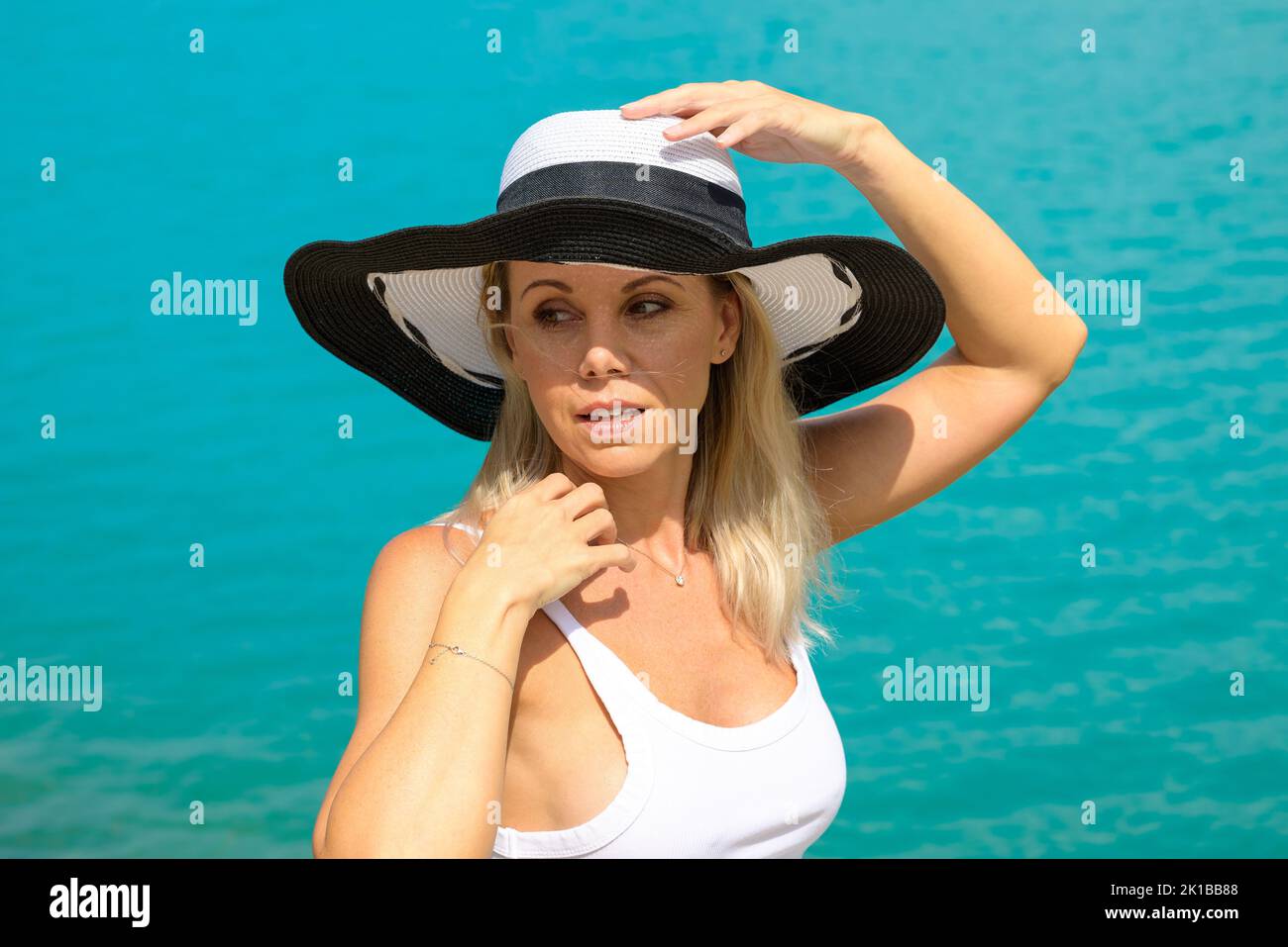 Attractive blond woman smiling friendly wearing a black and white summer hat in a white top with her hands on the hat in front of a turquoise sea Stock Photo