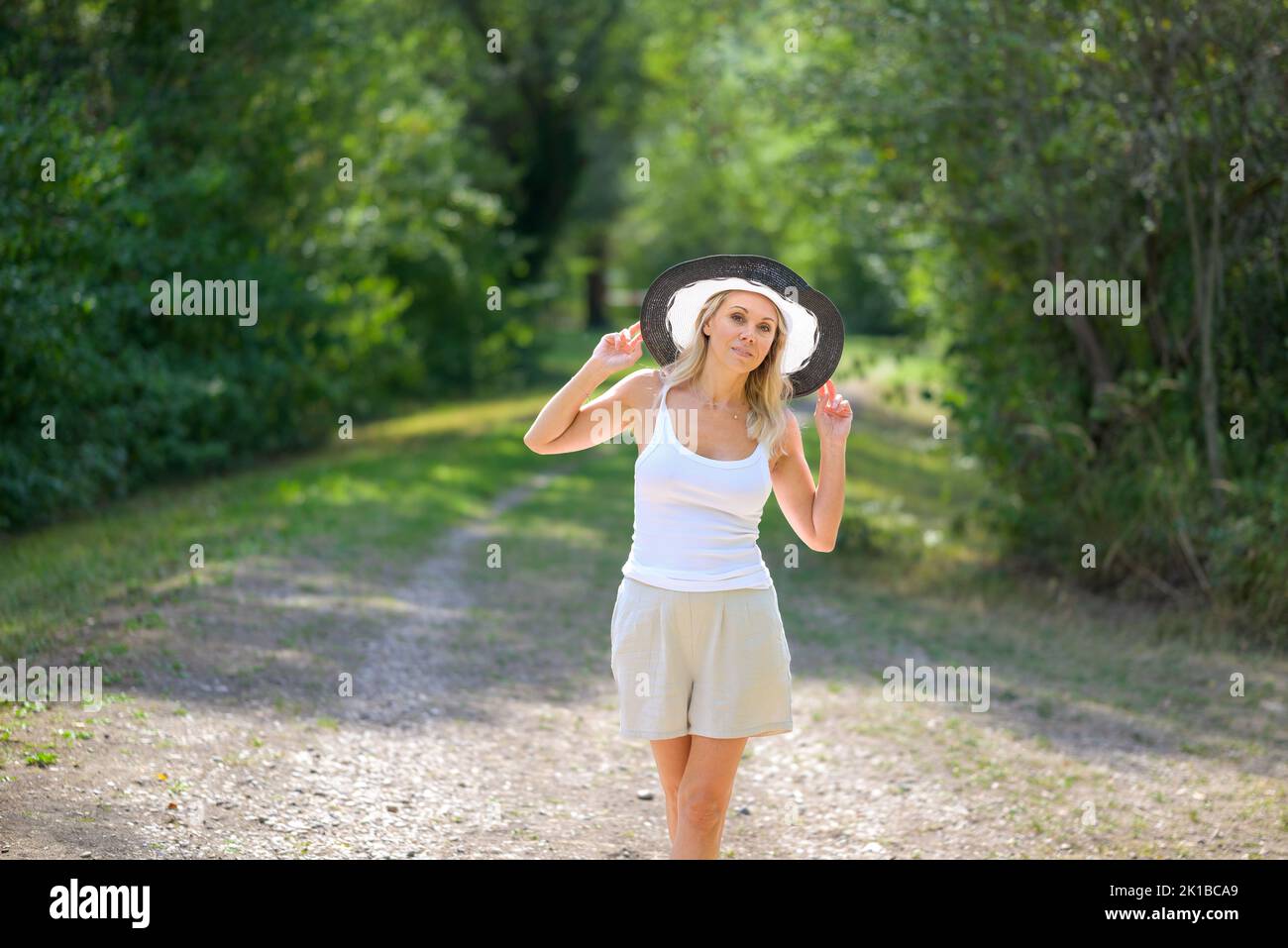 Attractive blond woman smiling friendly wearing a black and white summer hat in a white top with her hands on the hat in front of greenery Stock Photo