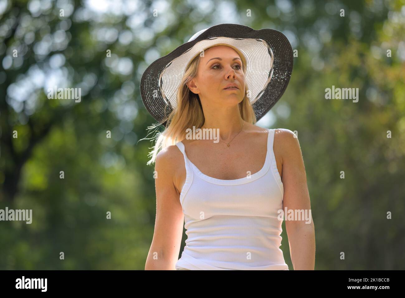 Attractive blonde woman wearing a black and white summer hat in a white top is walking along a path with green trees in the background looking aside Stock Photo