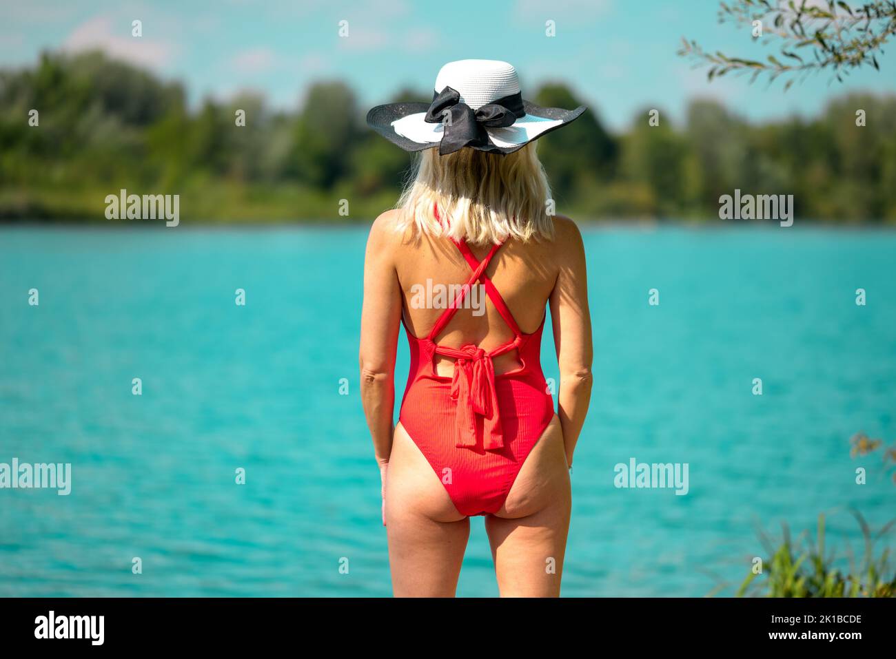 Rear view of a blonde woman with a black and white summer hat in a red swimsuit in front of a beautiful lake Stock Photo