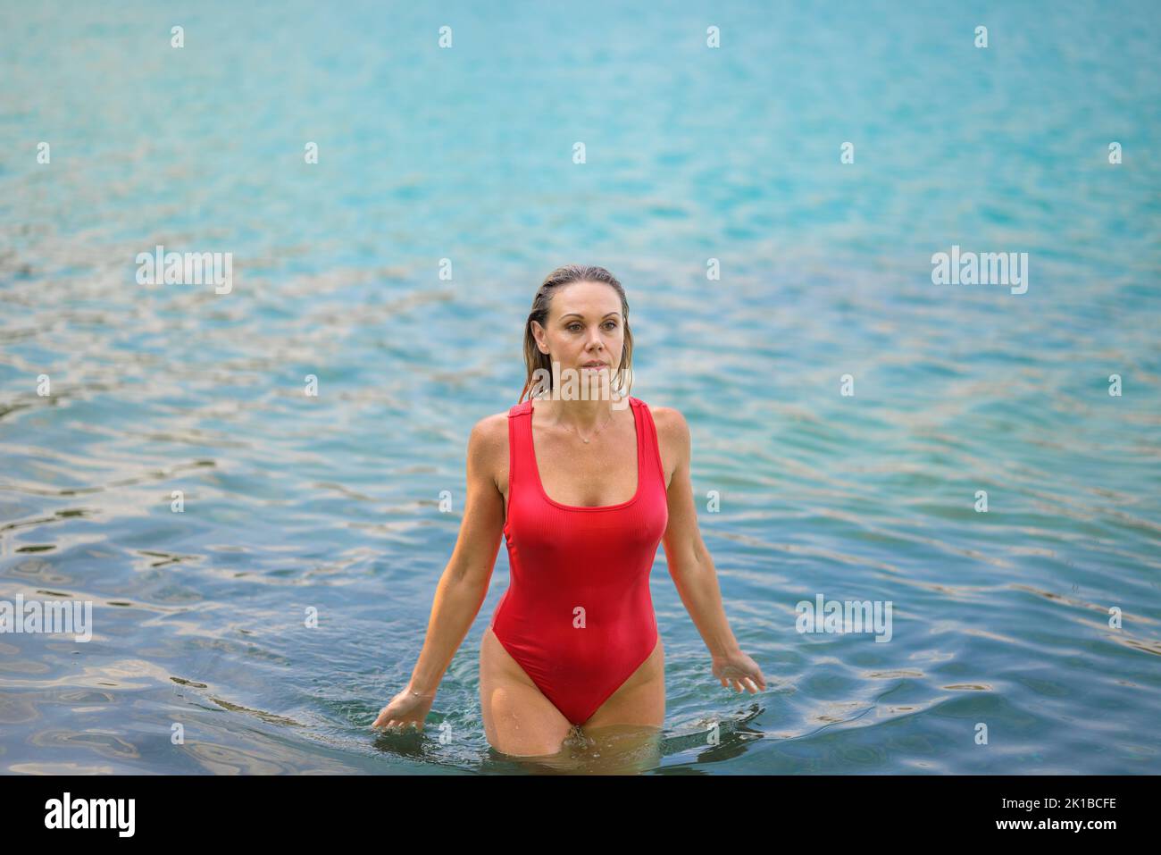 Gorgeous attractive blond woman with wet hair in a red swimsuit is slowly walking through the water deep in thought Stock Photo