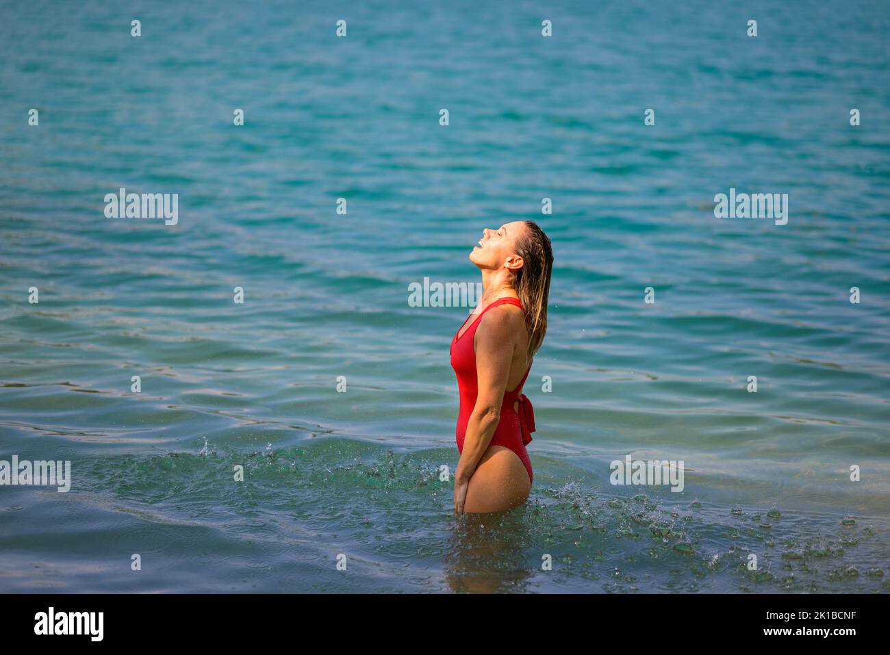 Gorgeous attractive blond woman with wet hair in a red swimsuit is standing in the water with closed eyes and enjoying the water Stock Photo