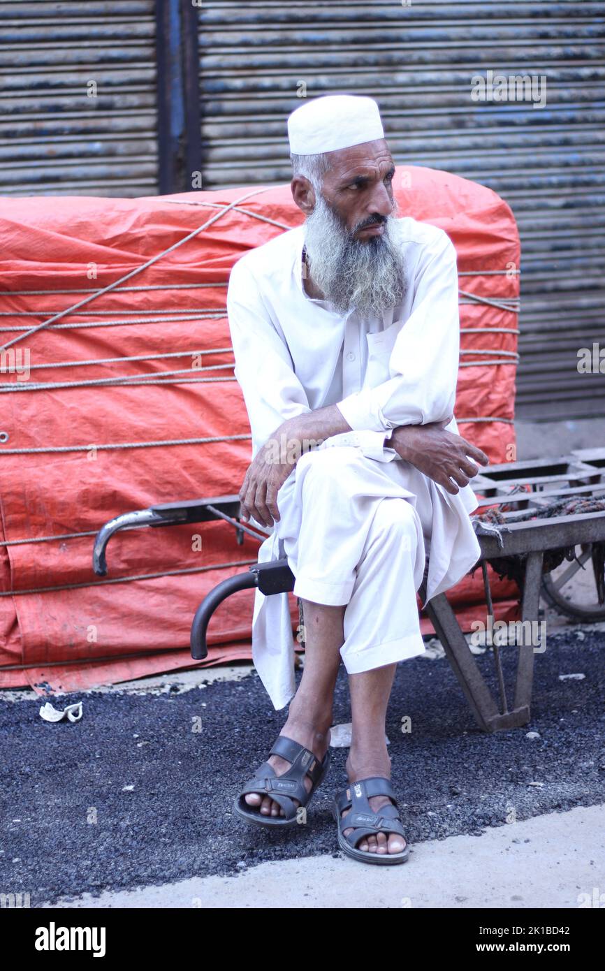A vertical shot of a Pakistani old male with a white beard in a white topi and kurta sitting in the street Stock Photo