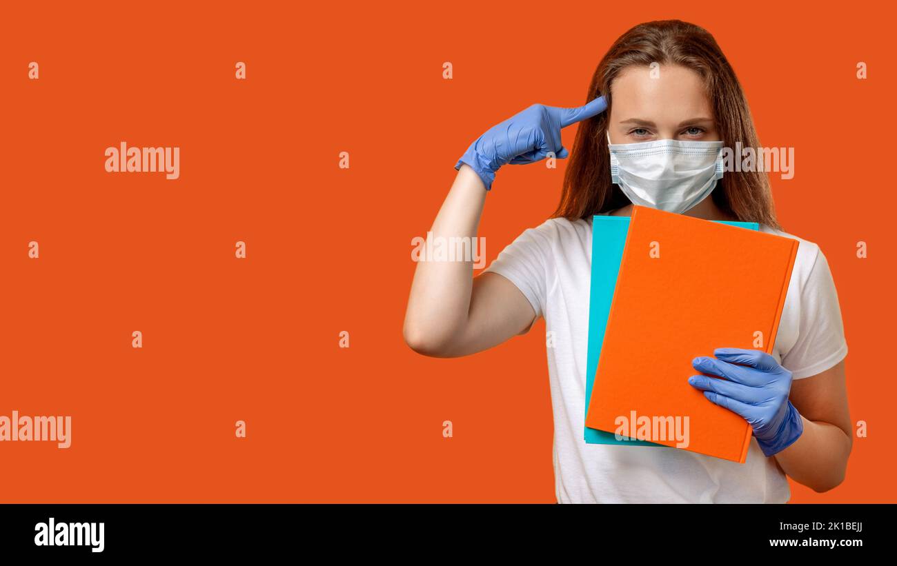 Quarantine education. Hygiene measures. Mindful female teacher in protective face mask gloves with books pointing at head isolated on orange red copy Stock Photo