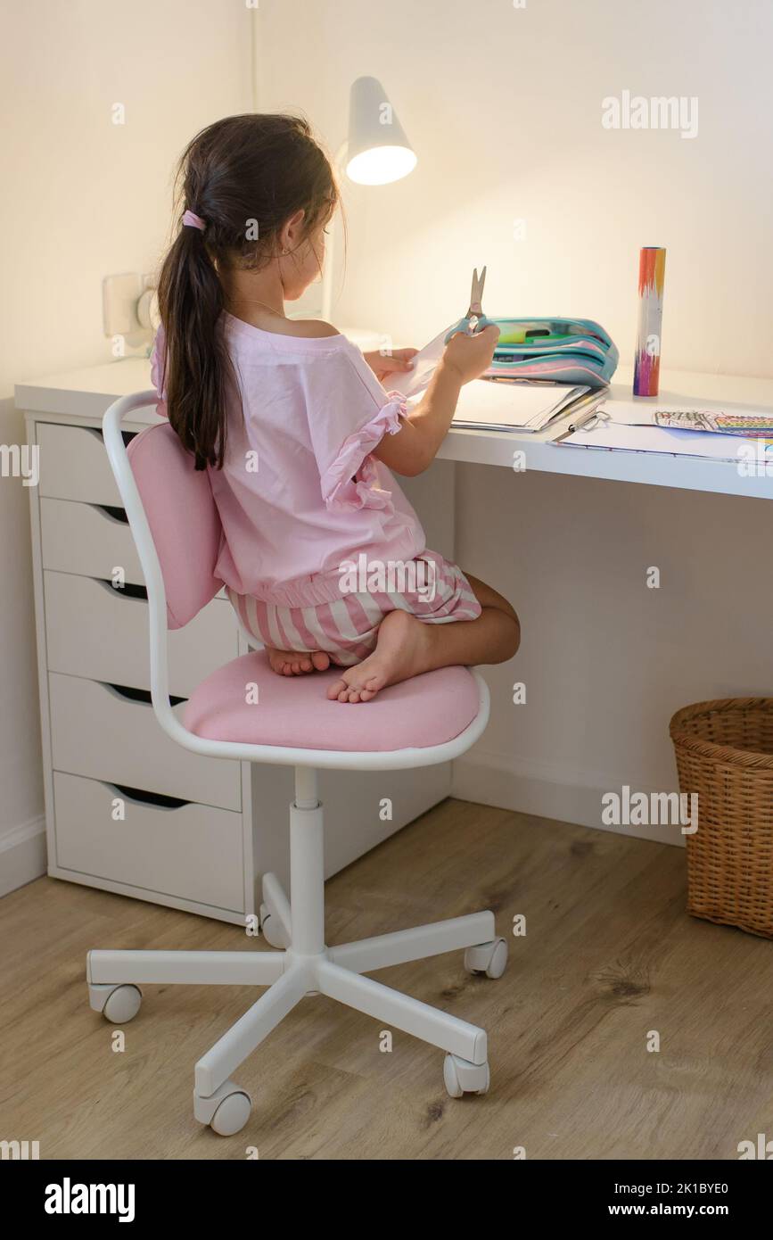 Adorable little girl with long dark hair in casual clothes sitting on pink student chair at white desk with scissors in hand in modern room. Stock Photo
