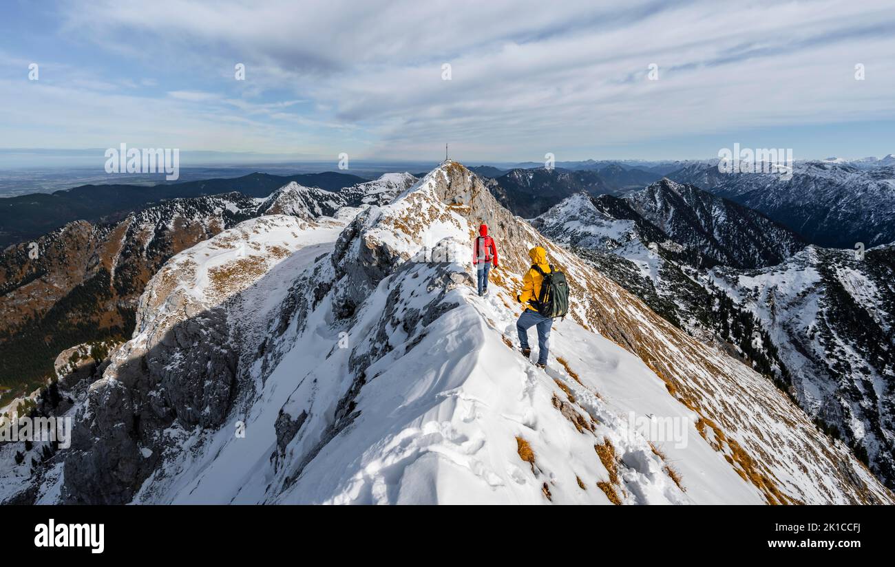 Two mountaineers on a narrow rocky snowy ridge, in the back summit of the Ammergauer Hochplatte with summit cross, view of mountain panorama, hiking Stock Photo
